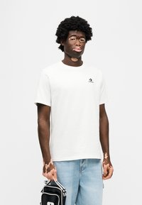 Λευκό βαμβακερό T-shirt με κοντά μανίκια και μικρό μαύρο λογότυπο στο στήθος. Το μοντέλο κρατάει μια ασπρόμαυρη τσάντα και φοράει ανοιχτόχρωμα μπλε τζιν.