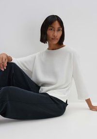 White ribbed sweater with a round neck and three-quarter sleeves, paired with dark loose-fitting pants. Smooth texture and relaxed fit.