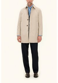 Beige waterproof trench coat with a classic collar, single-breasted button closure, side pockets, tailored fit, worn over a blue shirt and dark trousers.