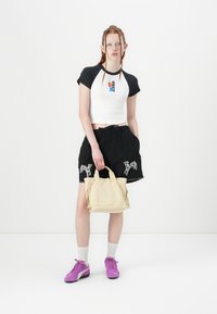 Balta ir juoda trumpa marškinėlių su grafiniu dizainu, juodos šortai su gyvūnų raštu, smėlio spalvos rankinė ir violetiniai sportbačiai.
