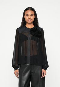 Blusa nera trasparente con maniche lunghe, design plissettato, colletto rotondo e un fiore decorativo in tessuto sulla spalla sinistra. Abbinata a pantaloni neri.