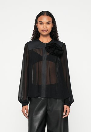 Sort gennemsigtig bluse med lange ærmer, plisseret design, rund krave og en dekorativ stofblomst på venstre skulder. Lægges sammen med sorte bukser.