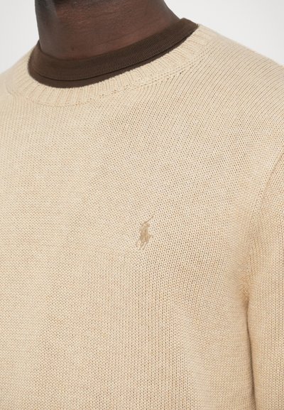 Béžový pletený sveter s okrúhlym výstrihom a hnedým rebrovaným golierom, na hrudi s malým vyšívaným logom značky.