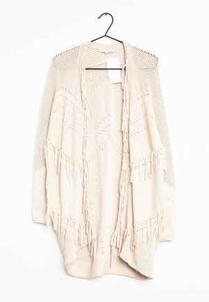 Cardigan beige en tricot avec ouverture sur le devant, motifs texturés et détails à franges, suspendu à un cintre noir sur fond blanc.
