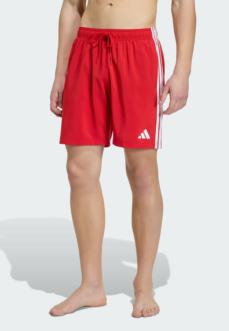 Oberkörperfreie Person trägt rote Adidas-Badeshorts mit weißen Streifen und Logo und steht barfuß auf einer hellen Oberfläche.