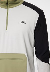 Lys grå pullover med olivengrønne accenter og sorte skuldre. Har en høj krave, frontlomme med lynlås og logo på brystet.