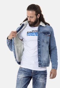 Jeansjacke mit Fleece-Futter, hellblaue Farbe, Knopfleiste vorne, Brusttasche und leichten Waschungseffekten; getragen über einem weißen T-Shirt mit Grafik.