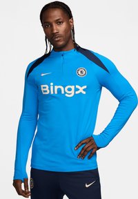 Maglia sportiva a maniche lunghe di un vivace blu, con colletto a mezza zip, accenti sulle spalle in blu navy e un logo dorato con testo sul davanti.
