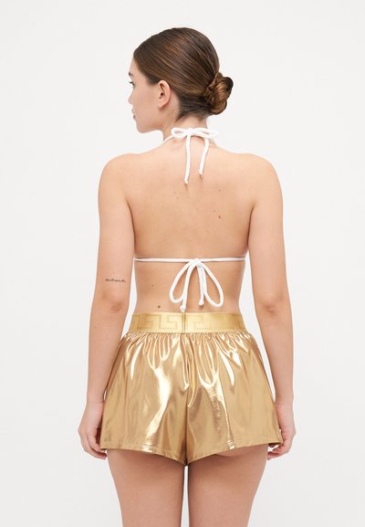 Haut de bikini blanc à col bateau avec un nœud dans le dos, associé à un short doré brillant avec un motif décoratif à la taille. Texture lisse dans l'ensemble.