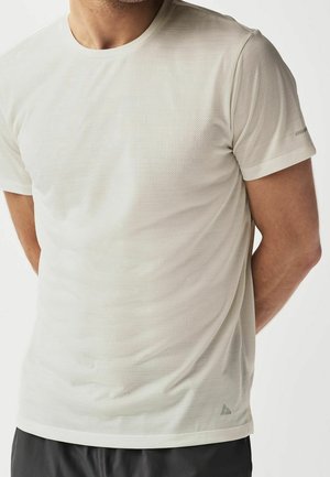 Homme portant un t-shirt texturé beige clair à manches courtes, debout avec les mains derrière le dos sur un fond blanc.