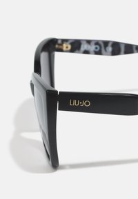 Black sunglasses with a wide rectangular frame. Gold logo on the temple, textured grey and black pattern on the inner arm. Smooth finish.