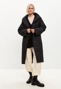 Sort pufferjakke med hætte, med knaplukning foran, brede ærmer og lang længde, båret over en lys sweater og cremefarvede bukser.