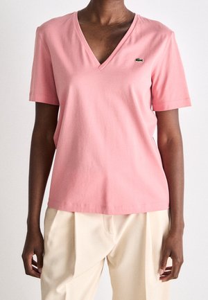 Osoba ubrana w różowy krótki rękaw z dekoltem w serek z małym zielonym logo krokodyla oraz jasnobeżowe plisowane spodnie.