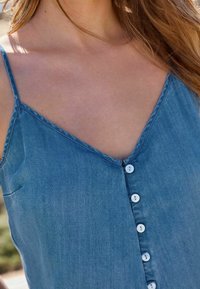 Zbliżenie na osobę ubraną w bezrękawnik z niebieskiego dżinsu z białymi guzikami z przodu, w naturalnym świetle.