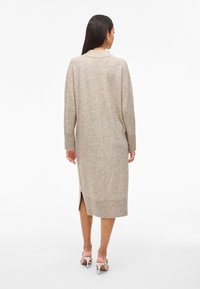 Bézs színű kötött ruhakötény hosszú ujjakkal, magas nyakkivágással, oldalsó hasítékokkal, bordázott mandzsettával és szegéllyel. Puha textúrájú és közép-vádli hosszúságú.