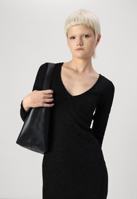 Vestido preto com mangas longas e decote em V, padrão texturizado. Mala de couro preta com design afunilado, segurada com uma mão.