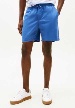 Bañador azul con cinturilla elástica y cordón ajustable, que cuenta con bolsillos laterales y un pequeño acento de logo. Combinado con zapatillas blancas.