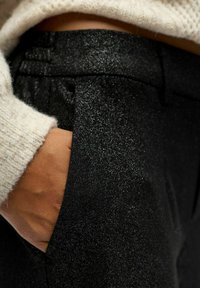 Schwarze Hose mit strukturierter, glänzender Oberfläche, ausgestattet mit einer Tasche und einem leicht schimmernden Finish, kombiniert mit einem hellen, flauschigen Pullover.