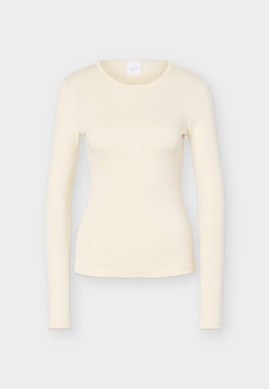 Krémovo sfarbená priliehavá košeľa s dlhými rukávmi a okrúhlym výstrihom, zobrazená na jednolivom svetlosivom pozadí.