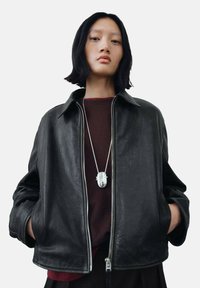 Black leather jacket featuring a zip closure, collar, and loose fit. Worn over a dark red top, with a silver pendant necklace as an accent.