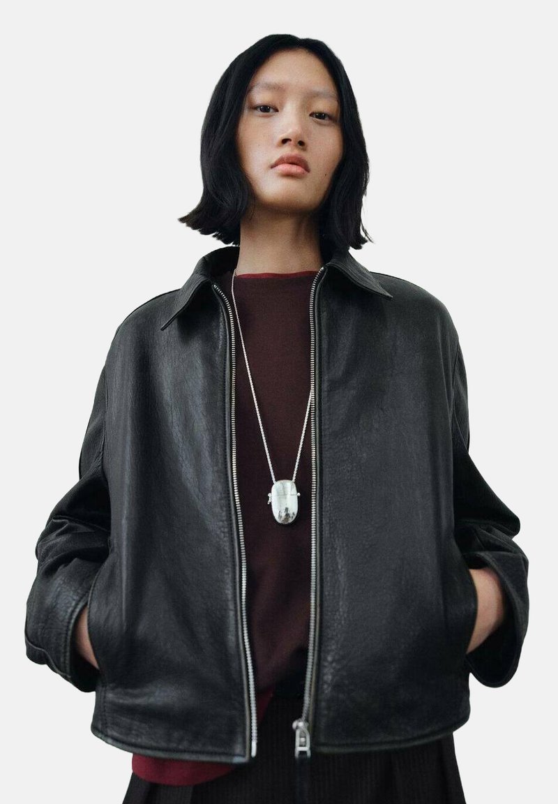 Black leather jacket featuring a zip closure, collar, and loose fit. Worn over a dark red top, with a silver pendant necklace as an accent.