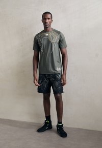 T-shirt vert olive avec un design graphique doré, associé à un short noir avec un motif audacieux. Baskets noires avec des accents néon.
