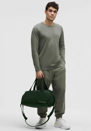 Top de manga larga en color oliva y pantalones jogger combinados con un bolso de deporte de color verde oscuro. El bolso cuenta con asas dobles y una correa de hombro desmontable.