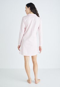 Nuisette à carreaux rose clair en tissu doux, avec des manches longues, un col et un ourlet arrondi. Détails de couture visibles.