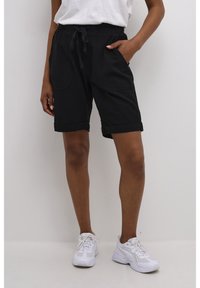 Pantalones cortos de algodón negros con un ajuste relajado, que cuentan con una cinturilla elástica y cordón, bolsillos laterales y dobladillos ajustados. Combinados con zapatillas blancas.
