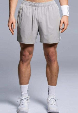 Mann trägt hellgraue Sportshorts, weiße Sportsocken, weiße Turnschuhe und ein weißes Armband am rechten Handgelenk, steht auf grauem Boden.