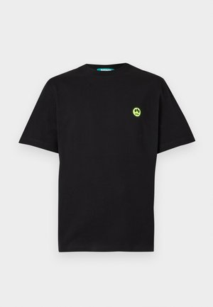 Mladý muž stojaci s rukami v vreckách, oblečený v čiernom tričku a teplákoch so žltými smajlíkmi a bielymi teniskami.