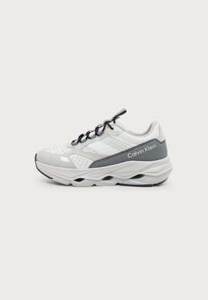 Hvid og grå Calvin Klein sneaker med sorte snørebånd og en tyk, skulptureret sål, vist fra siden på en ensfarvet baggrund.