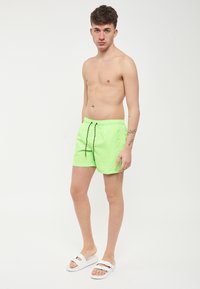Pantaloni da bagno verde neon con coulisse nera, caratterizzati da una texture liscia e una vita elastica, abbinati a sandali bianchi.