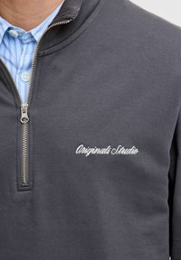 Sudadera de cuello alto gris con textura suave, cierre zip plateado y bordado "Originals Studio" en blanco en la parte izquierda del pecho.
