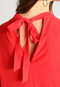 Röd blus med V-formad rygg, nyckelhålsöppning och knytning i nacken. Tillverkad av lätt, strukturerad tyg.