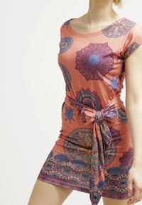 Femme portant une robe corail à manches courtes avec des motifs mandala colorés et une ceinture en tissu nouée autour de la taille.