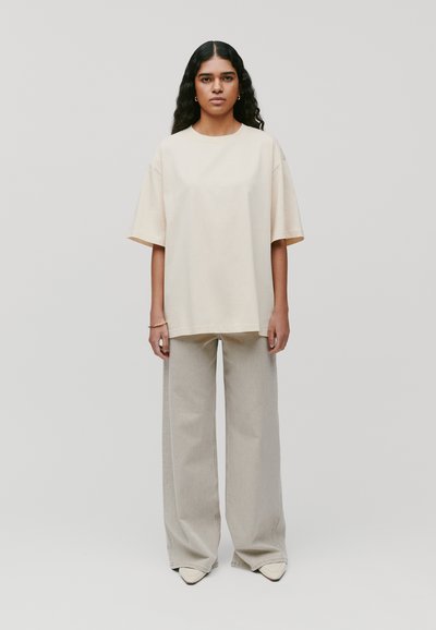 Beżowa oversize koszulka z krótkim rękawem, zestawiona z jasnoszarymi spodniami w szerokim kroju i czółenkami w odcieniu kremowym. Prosty i minimalistyczny design.