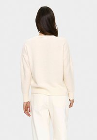 Femme aux longs cheveux sombres portant un pull couleur crème et un pantalon blanc cassé, se tenant de dos à la caméra devant un fond uni.