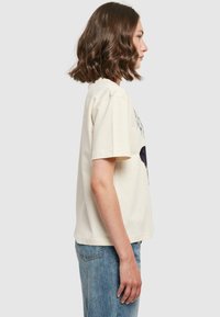 Cremefarvet t-shirt med korte ærmer, prydet med en trykt sort grafik på fronten. Casual pasform med rund halsudskæring, parret med lyse denimjeans.