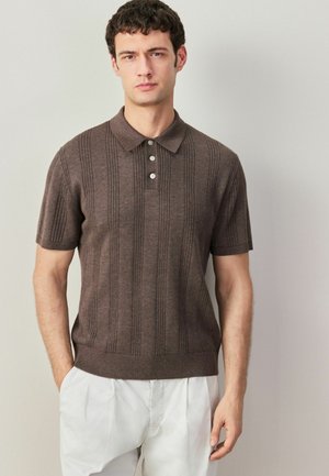 Brauner kurzärmeliger Polo-Pullover mit vertikaler Rippstruktur und Drei-Knopf-Leiste, kombiniert mit weißen Hosen.