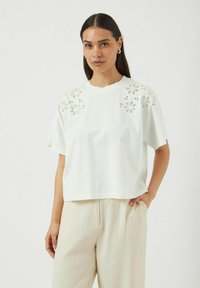 Hvit kortermet skjorte med blomsterbroderi i øyet på skuldrene, laget av et mykt stoff, med rund hals og løs passform.