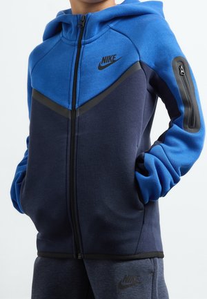 Niño con sudadera con capucha Nike azul y azul marino con bolsillo con cremallera en el brazo y pantalones deportivos Nike azul marino a juego, con las manos en los bolsillos.