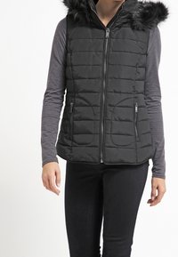 Černá prošívaná vesta s faux kožešinovou kapucí, zapínáním na zip a dvěma bočními kapsami. Nalezena na dlouhém šedém topu a černých kalhotách.