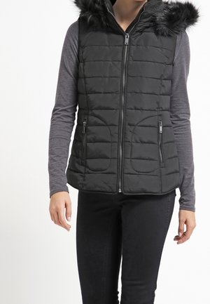 Sort, quiltet vest med en faux fur hætte, lynlås foran og to sidelommer. Båret over en langærmet grå top og sorte bukser.