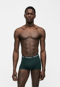 Άνδρας μοντέλο με σκουρόχρωμο δέρμα, κοντά μαλλιά και μυώδες σώμα, φοράει σκούρα πράσινα ανδρικά μποξεράκια Björn Borg, στέκεται σε απλό φόντο.