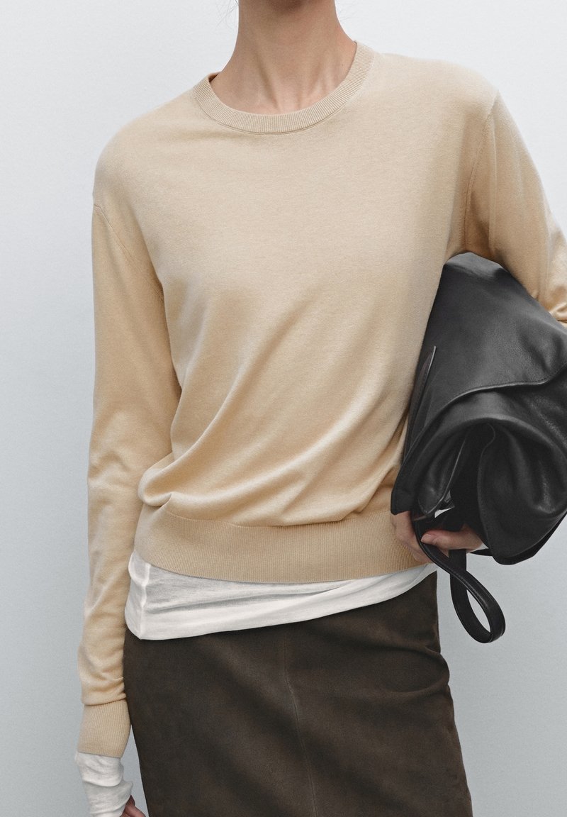 Personne portant un pull beige à manches longues sur une chemise blanche et un pantalon foncé, tenant un grand sac en cuir noir plié sous un bras.