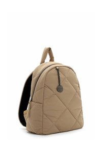 Bēša izšūta mugursoma ar melnu rāvējslēdzēju, augšējo rokturi, polsterētām plecu siksnām un apaļu metāla uzlīmi, kas karājas no rāvējslēdzēja roktura.