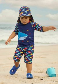 Set da bagno composto da una maglietta blu navy con una grafica di squalo e pesci multicolori, abbinata a pantaloni corti a fantasia. Le scarpe blu completano il look.