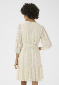 Vestido beige claro con mangas transparentes y abullonadas, escote con volantes y dobladillo en capas. Decorado con lentejuelas plateadas en un patrón de puntos.