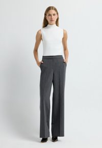 Top blanco sin mangas con cuello alto, combinado con pantalones gris de cintura alta. Los pantalones tienen un ajuste entallado y dos bolsillos laterales. Zapatos negros.